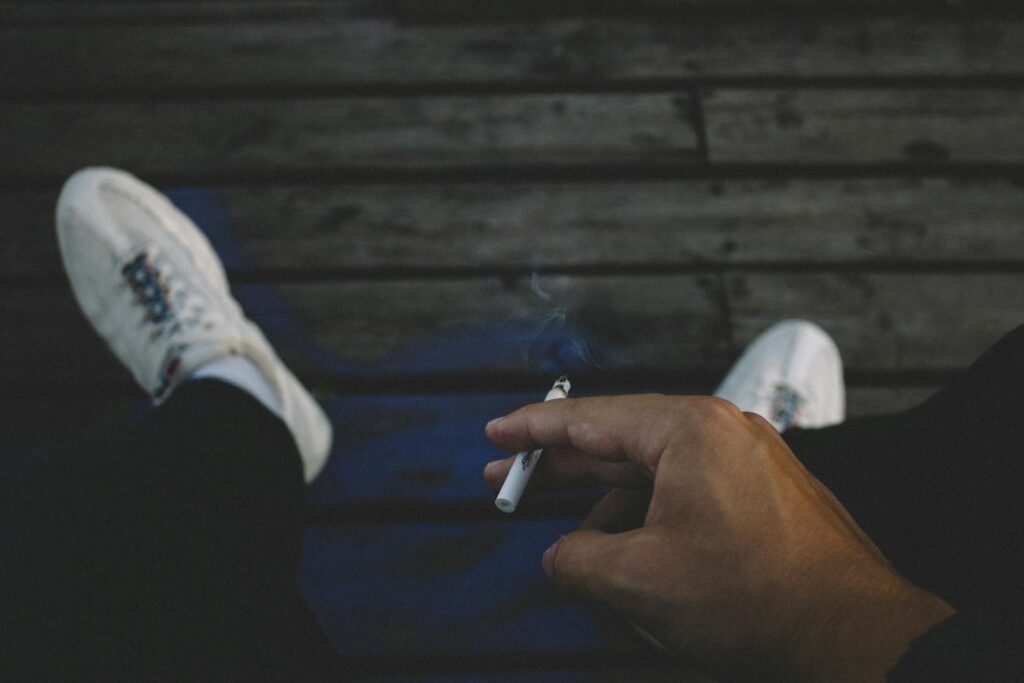 A man casually smoking a cigarette while relaxing outdoors on a wooden deck.